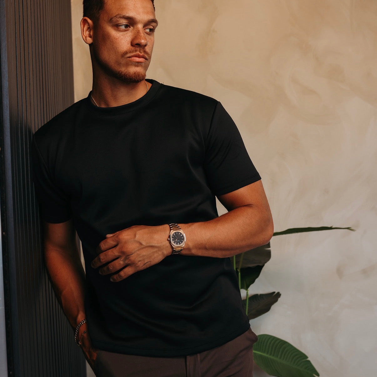 Model in het zwarte Gloss Essential Shirt, leunend tegen een donkere wand