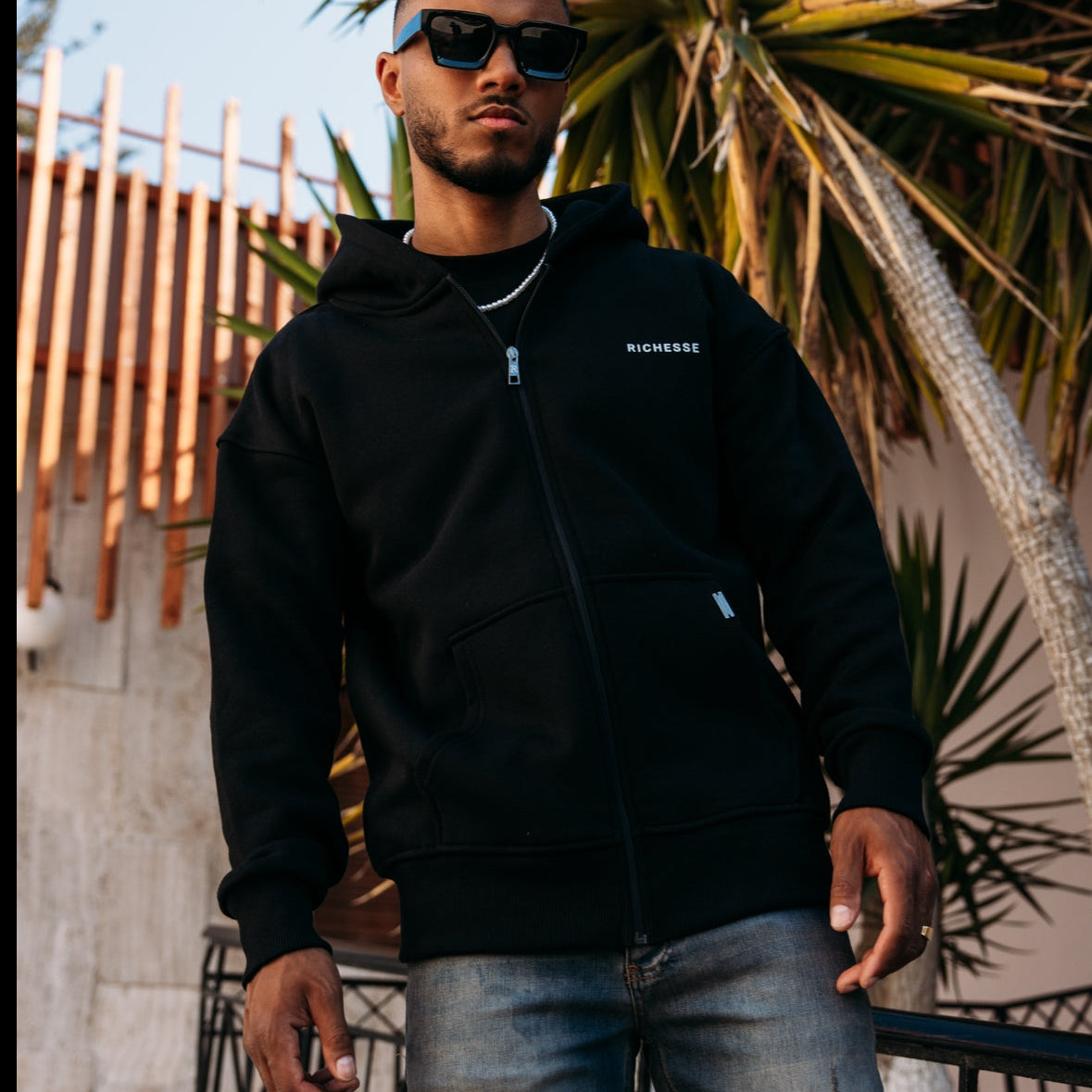 Model met zonnebril in een zwarte Richesse zip hoodie en lichte jeans, buiten tussen de palmbladeren