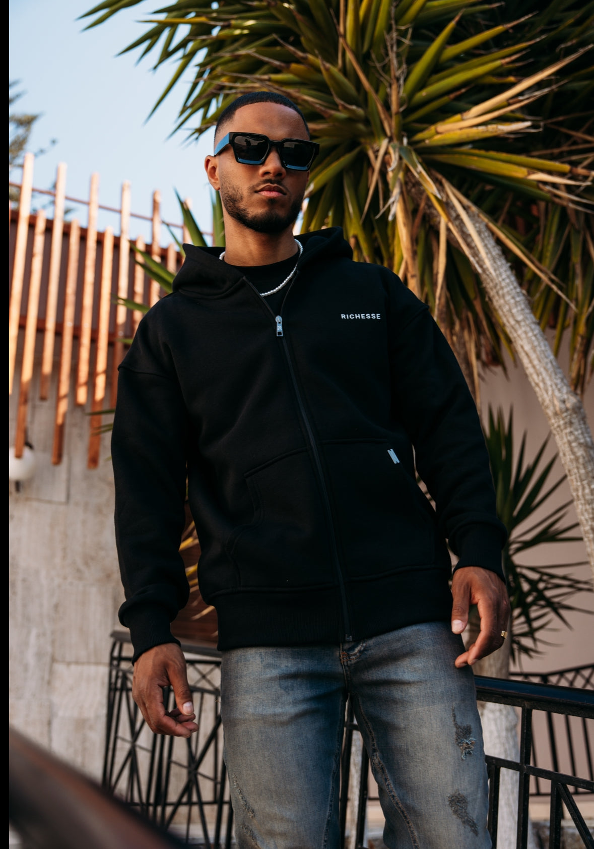 Model met zonnebril in een zwarte Richesse zip hoodie en lichte jeans, buiten tussen de palmbladeren