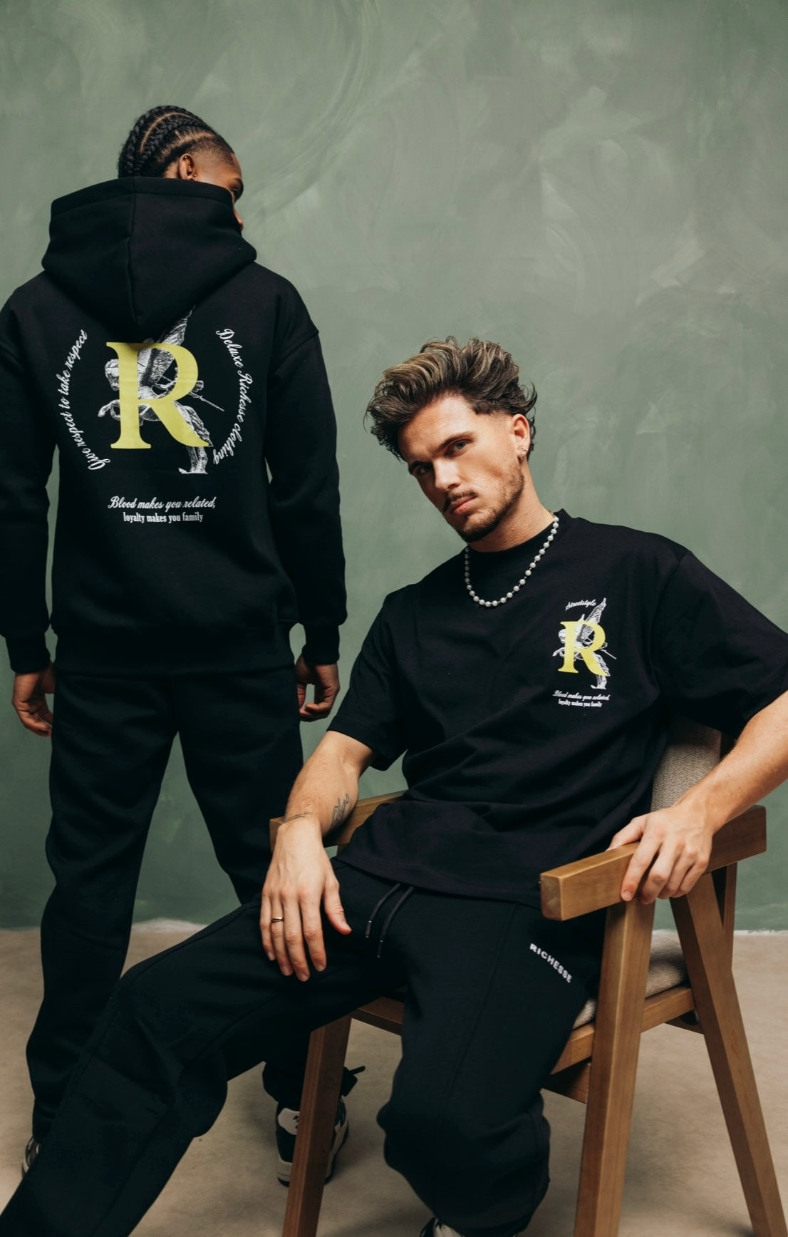 Twee modellen in zwarte Richesse-kleding met een groot R-logo, tegen een groene muur