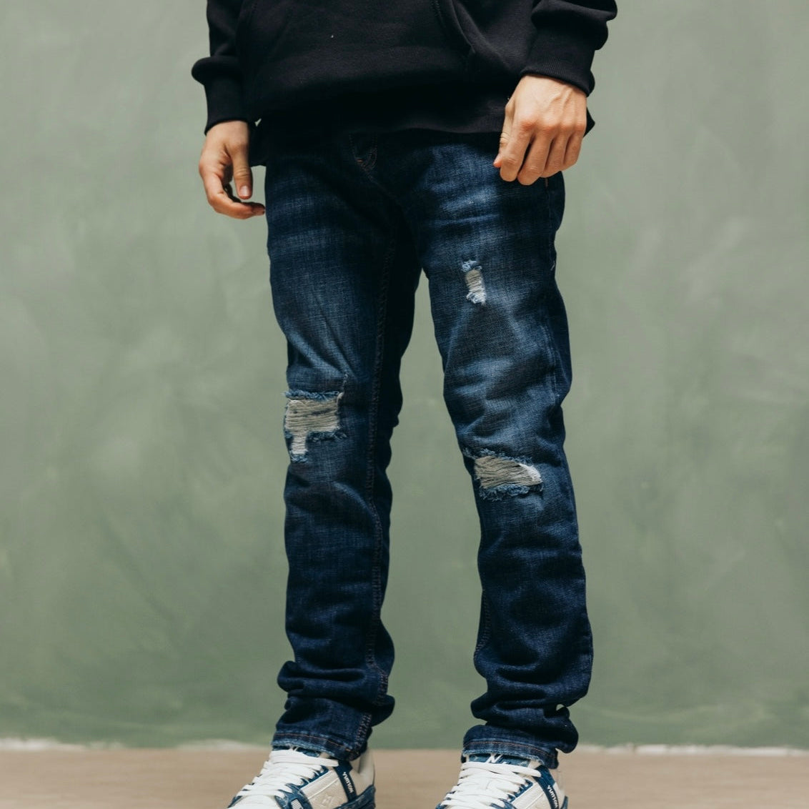 Model in de blauwe Prato Blue Jeans met sneakers, tegen een groene muur