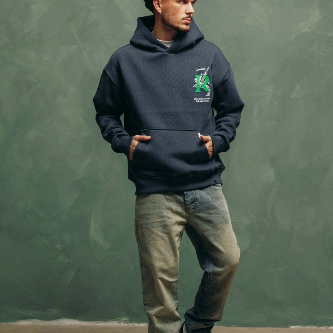 Model in de Artemis Hoodie met lichte jeans en sneakers, tegen een groene muur