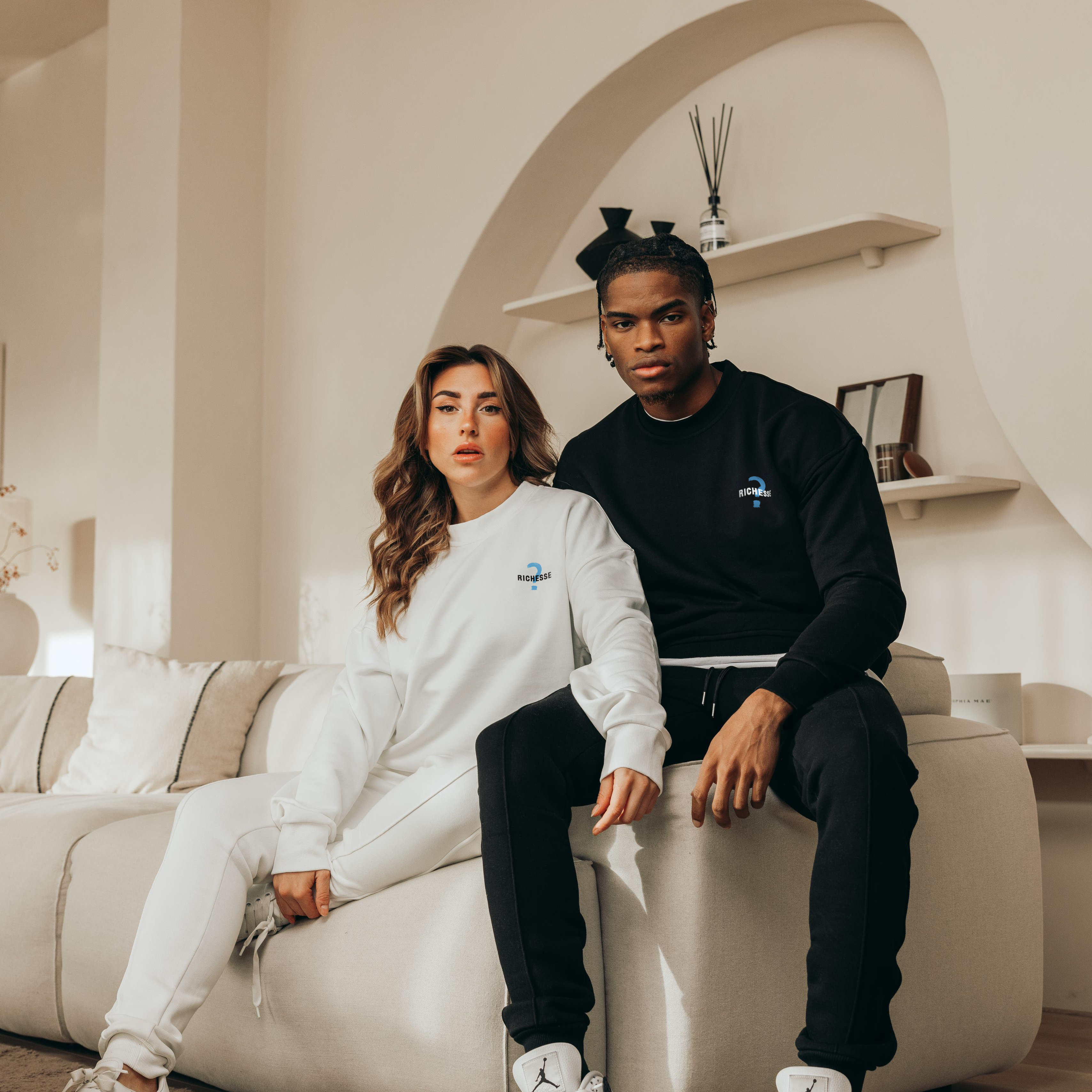 Een vrouw en een man in Richesse sweaters op een witte bank in een lichte kamer