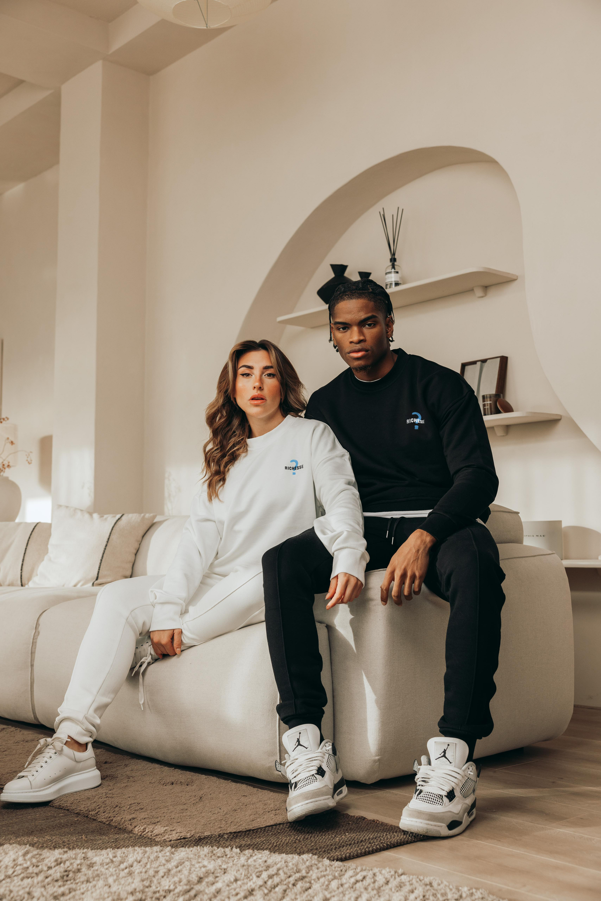 Een vrouw en een man in Richesse sweaters op een witte bank in een lichte kamer