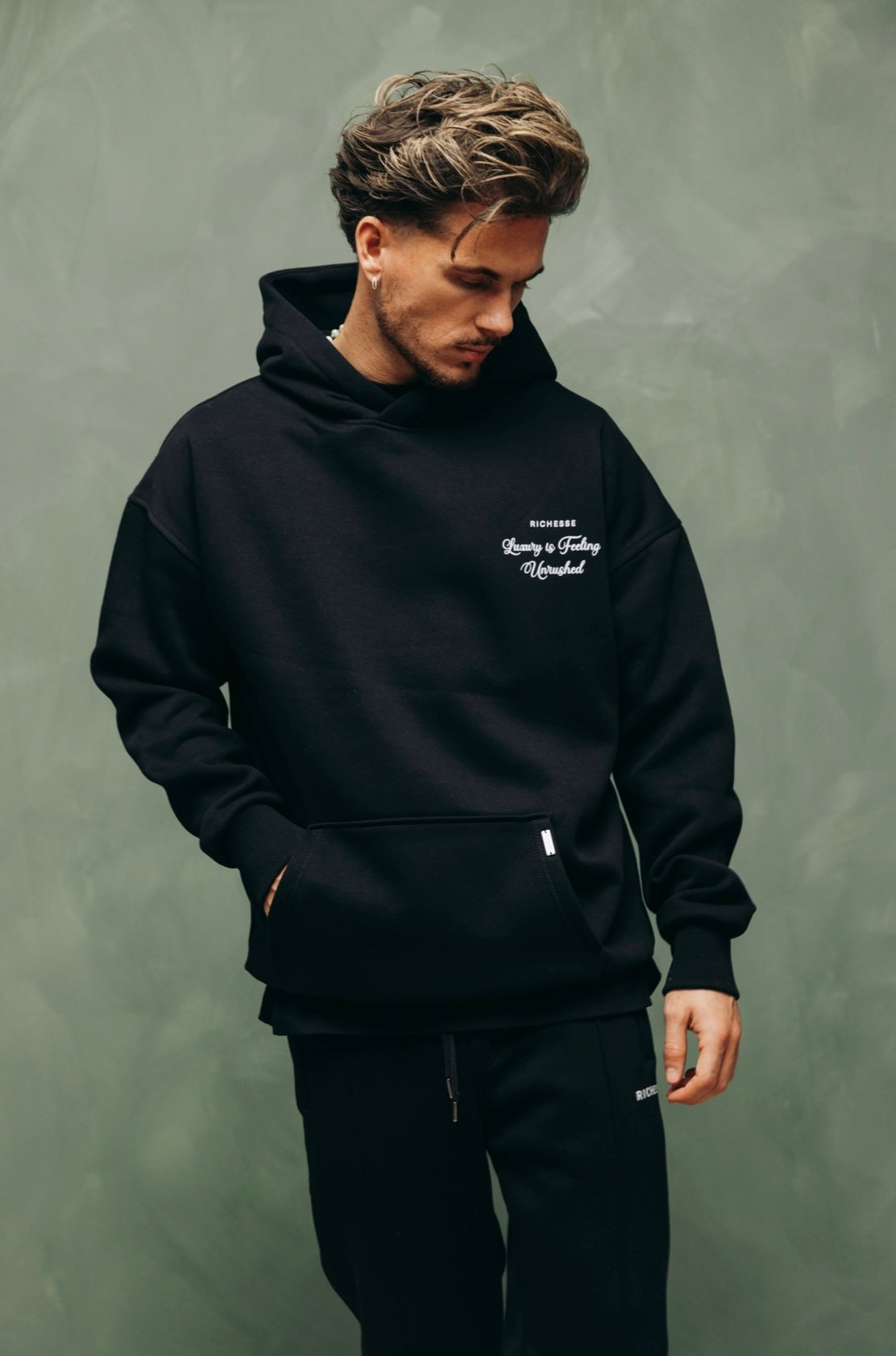Model in de zwarte Unrushed Hoodie met print op de borst, tegen een groene muur