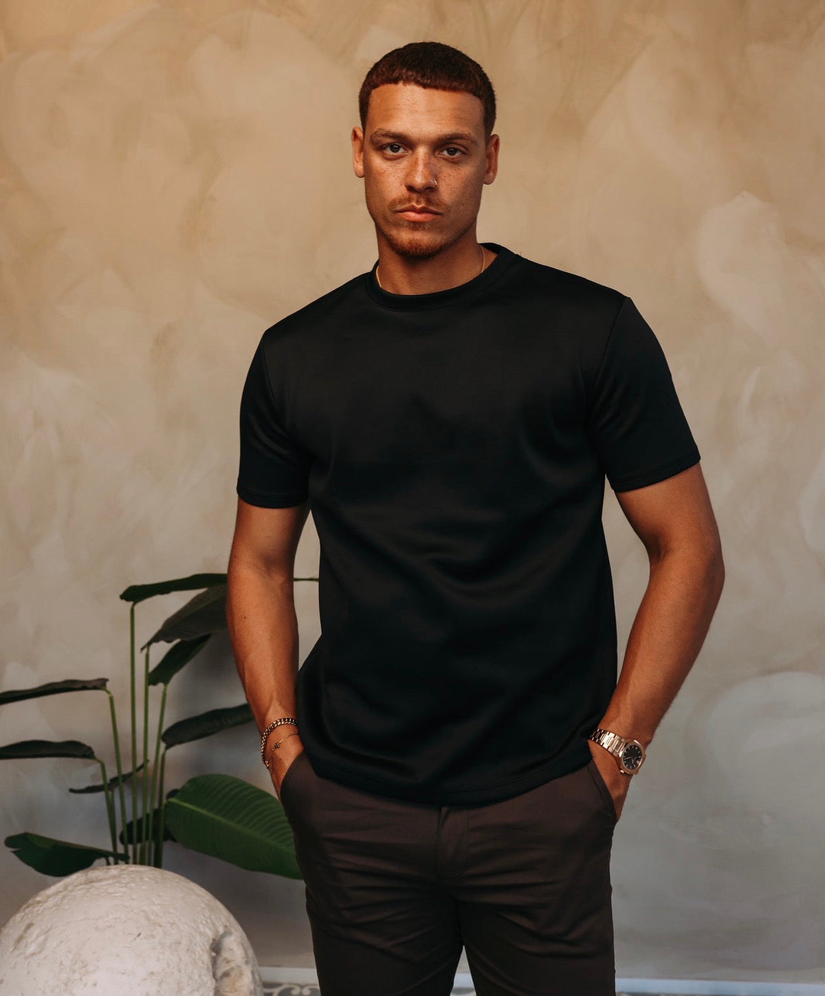 Model in het zwarte Gloss Essential Shirt met een donkere broek, tussen de planten