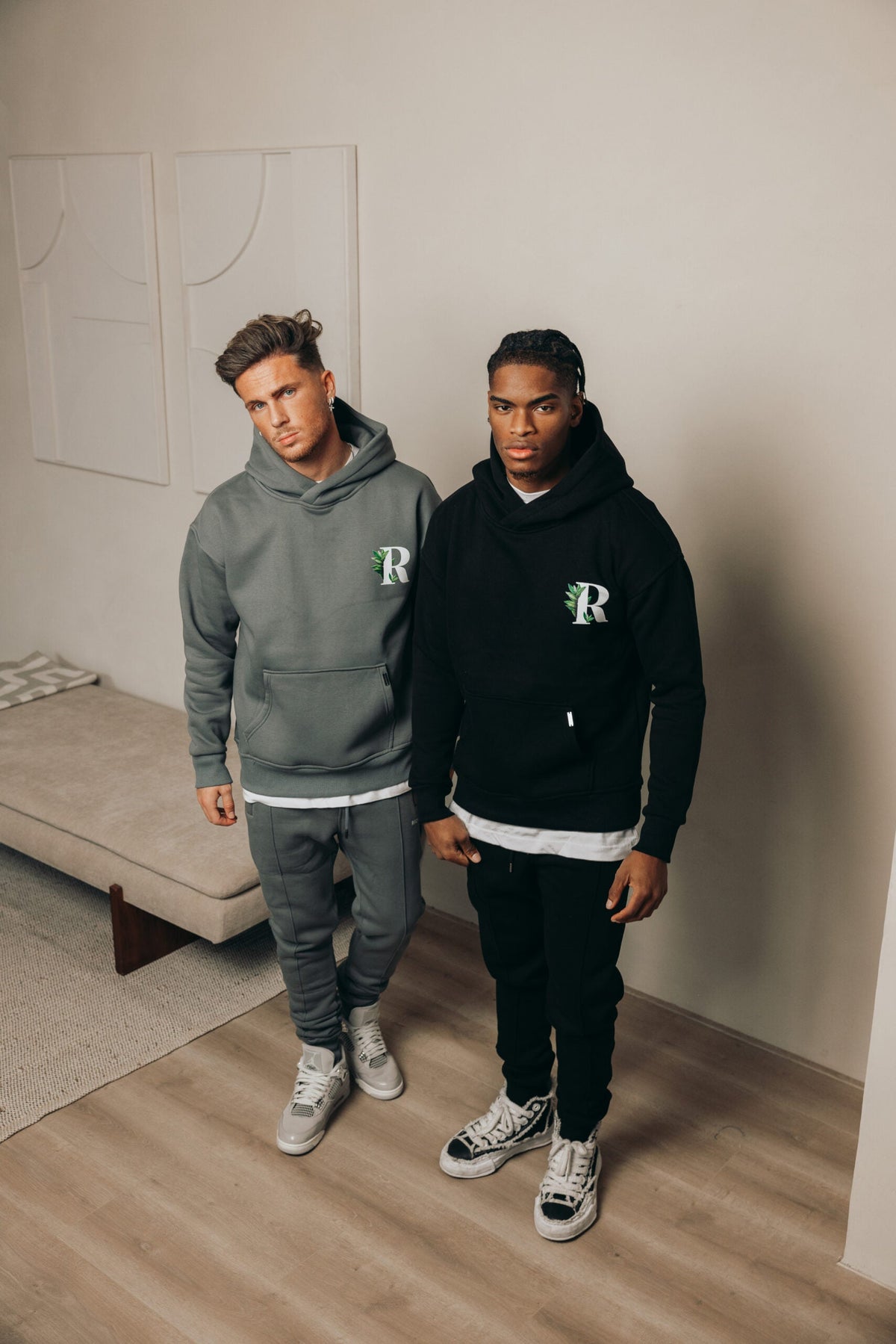 Twee modellen in een grijze en een zwarte Richesse hoodie met R-logo in een lichte kamer